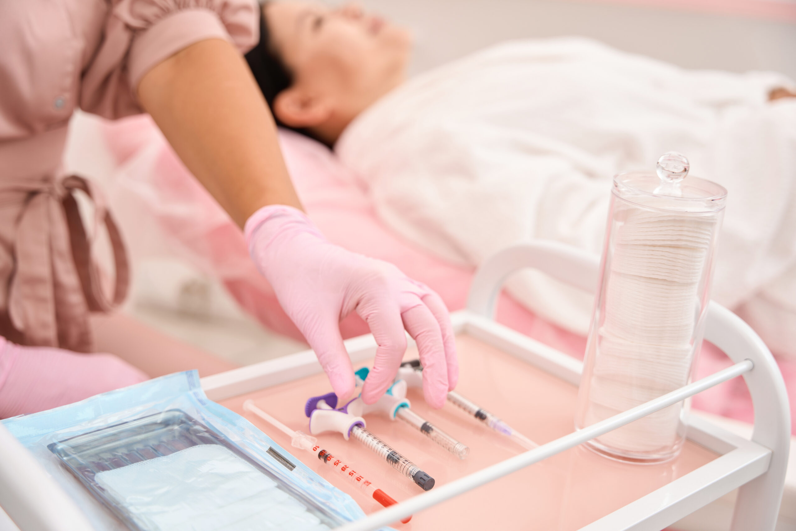 Woman in a pink uniform takes a syringe from an injection table, the client sits in a cosmetology chair