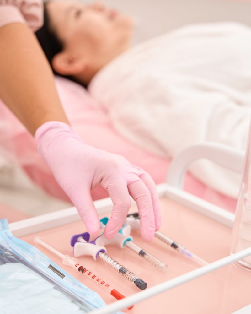 Woman in a pink uniform takes a syringe from an injection table, the client sits in a cosmetology chair